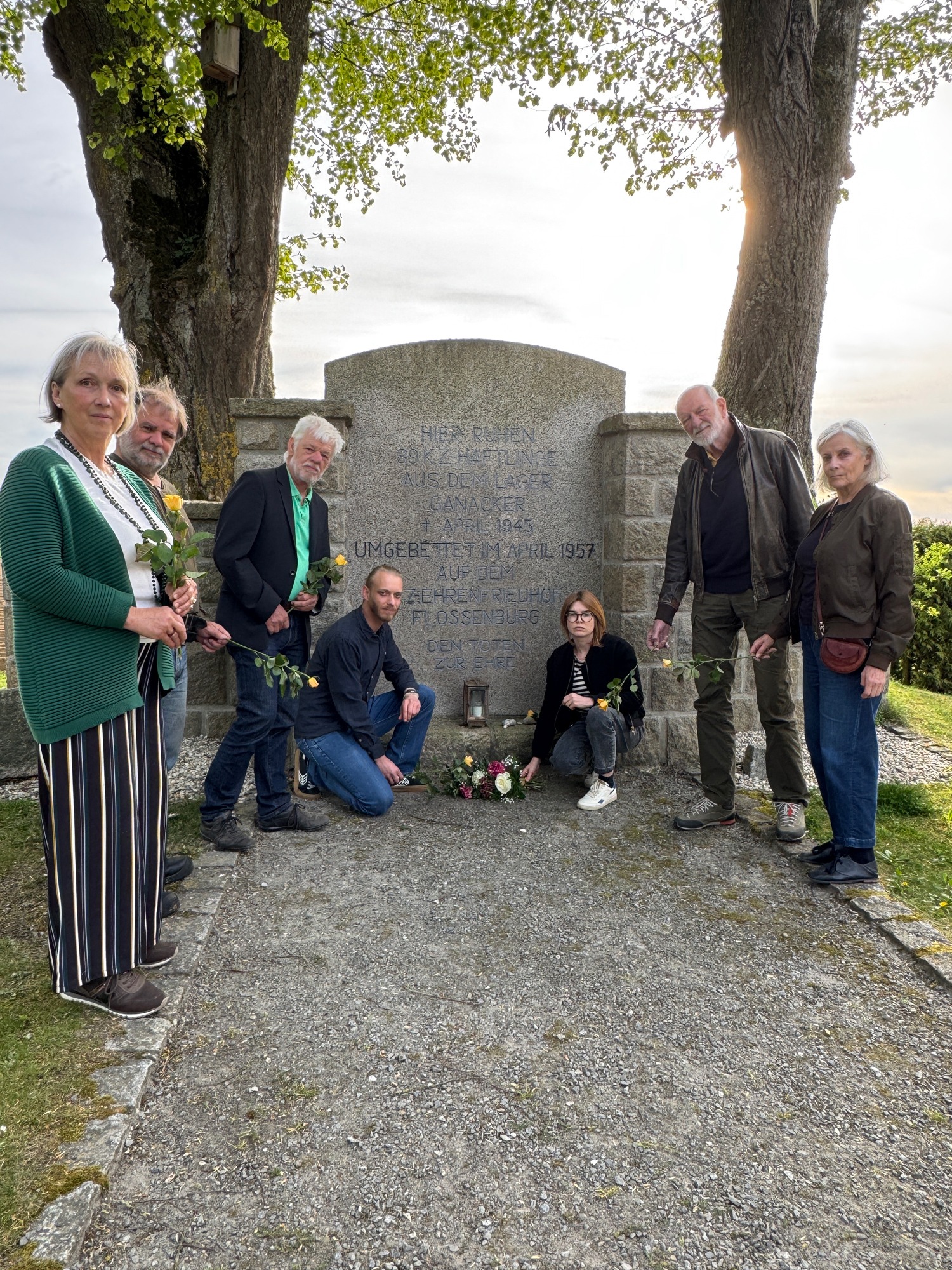 Eine große Gedenktafel steht in der Mitte und links und rechts davon eine Linde. 4 Männer und 3 Frauen legen BLumen Nieder und blicken in die Kamera.