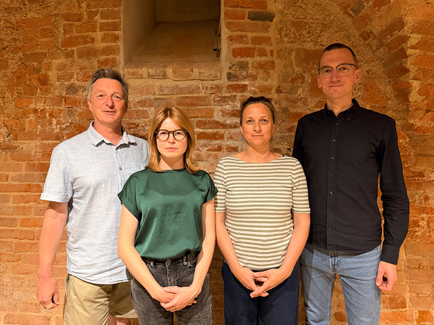 Gruppenfoto von vier Personen – zwei Frauen und zwei Männern – die nebeneinander vor einer rustikalen Backsteinwand stehen und freundlich in die Kamera blicken.