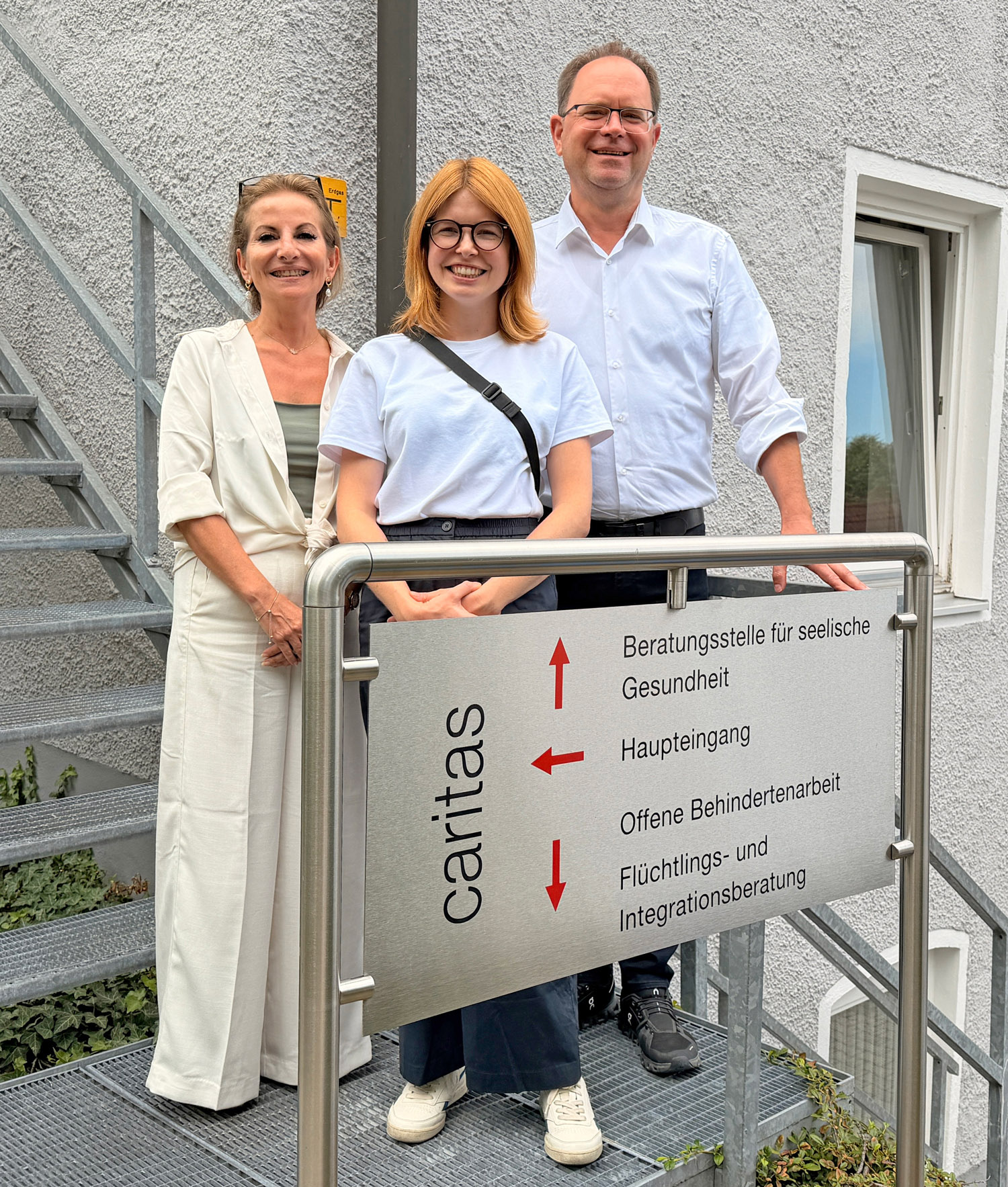 Drei Personen stehen lächelnd vor einem Caritas-Gebäude an einer Außentreppe, dahinter ein Hinweisschild mit den Aufschriften 'Beratungsstelle für seelische Gesundheit', 'Haupteingang', 'Offene Behindertenarbeit' und Flüchtlings- und Integrationsberatung.