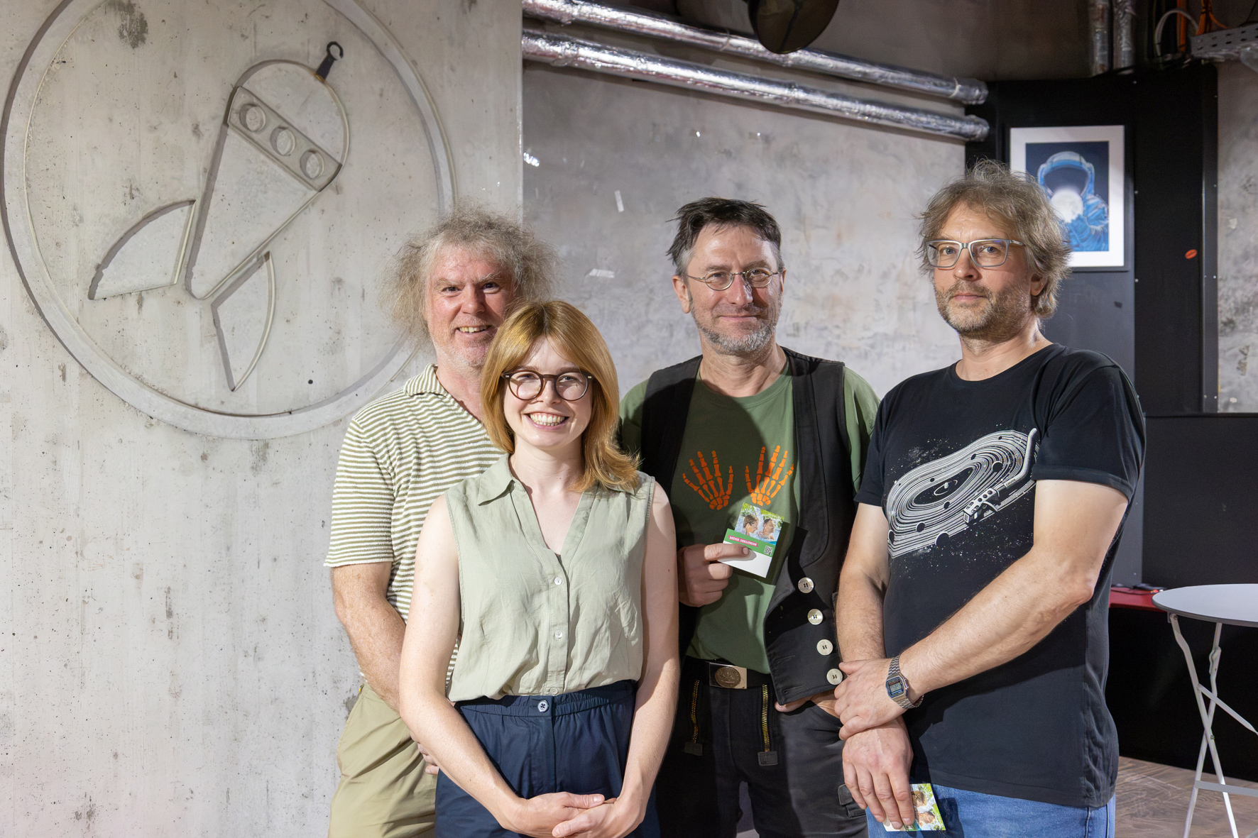 Gruppenfoto von vier Personen vor einer grauen Betonwand mit einem kreisförmigen Raketen-Logo. Von links: ein Mann mit grau-lockigem Haar im gestreiften Poloshirt, eine Frau mit schulterlangem Haar und Brille in hellgrüner Bluse, ein Mann mit Brille und Weste, der einen Gutschein in der Hand hält, sowie ein Mann mit Brille in dunklem T-Shirt mit Vinyl-Print. Alle lächeln in die Kamera.