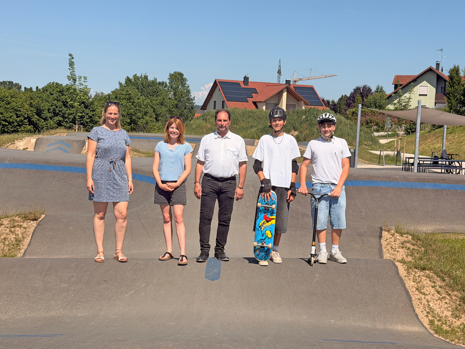 Fünf Personen stehen auf einem Betonparcours eines Pumptracks unter blauem Himmel. Eine Frau im blauen Kleid, eine Jugendliche in Shorts, ein Mann im weißen Hemd sowie zwei Jugendliche mit Fahrradhelmen und Skateboard bzw. Tretroller. Im Hintergrund Wohnhäuser mit Solaranlagen und ein Baukran.