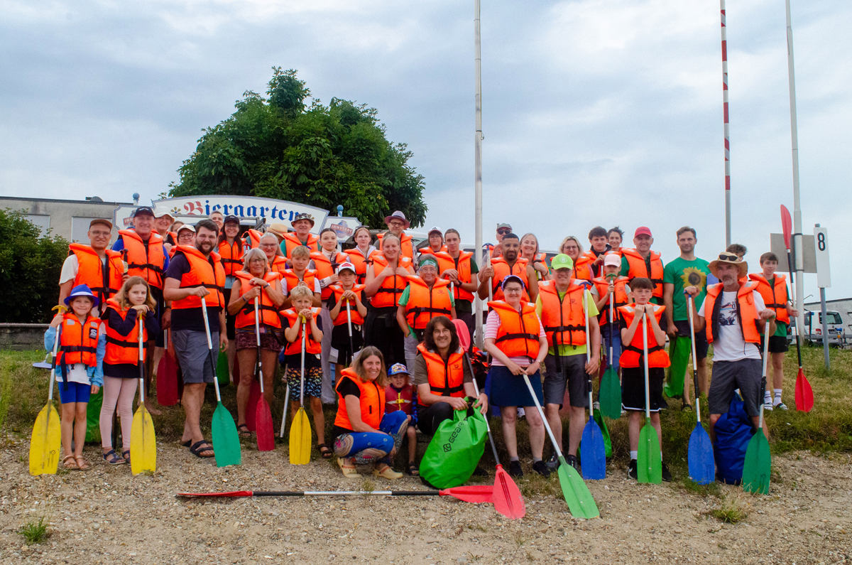 Große Gruppe von Erwachsenen und Kindern in orangefarbenen Schwimmwesten mit bunten Paddeln, stehen und knien gemeinsam für ein Gruppenfoto im Freien vor einem Biergarten-Schild, im Hintergrund Bäume und bewölkter Himmel.