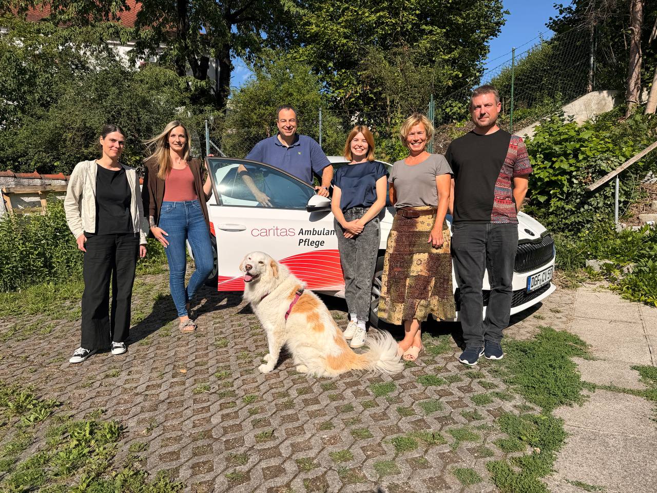 Sechs Personen stehen gemeinsam vor einem weißen Auto mit der Aufschrift 'Caritas Ambulante Pflege', im Vordergrund sitzt ein großer weiß-brauner Hund. Die Gruppe steht auf einem Pflasterweg vor einem grünen Gartenhintergrund bei Sonnenschein.