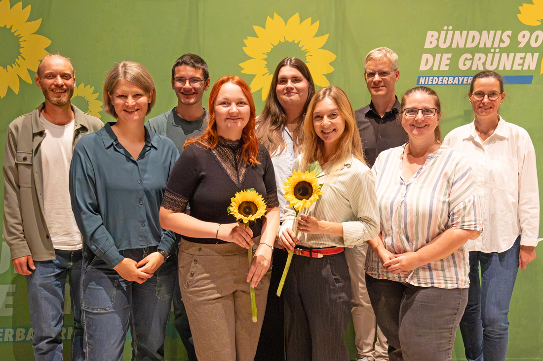 Eine Gruppe von neun Frauen und Männern steht lächelnd vor einer grünen Wand mit dem Logo von Bündnis 90/Die Grünen und gelben Sonnenblumen. Zwei der Frauen im Vordergrund halten jeweils eine Sonnenblume in der Hand.