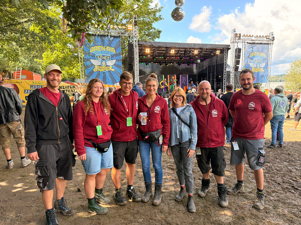 Eine Gruppe von acht Personen, überwiegend in weinroten Team-Shirts oder Hoodies, steht lächelnd nebeneinander auf einem Festivalgelände. Im Hintergrund ist eine große Open-Air-Bühne mit dem Schriftzug „Open-Air Steinberg“ auf blauen Bannern zu sehen. Das Wetter ist sonnig, und im Vordergrund ist der Boden unbefestigt und teilweise ausgetreten.