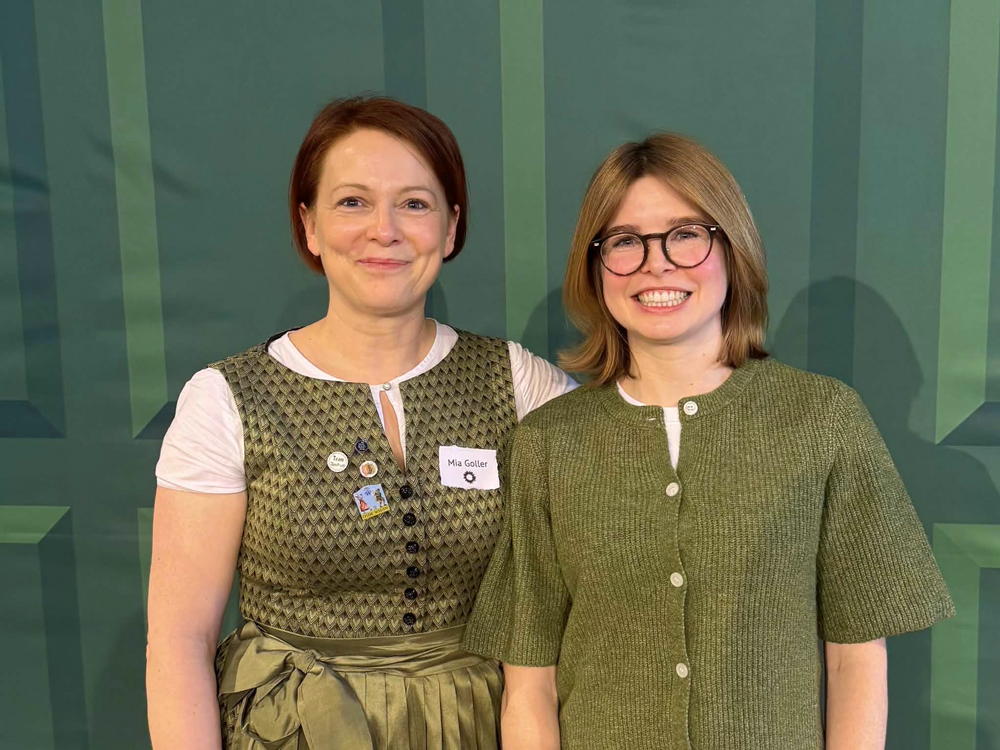 Porträt von zwei lächelnden Frauen vor einem dunkelgrünen Hintergrund. Die linke Frau trägt eine traditionelle grün gemusterte Weste mit Dirndlrock, die rechte Frau eine grüne Strickjacke und eine Brille.