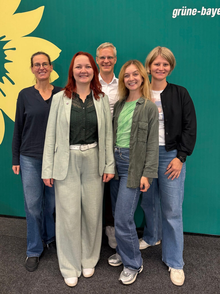  Fünf Personen – vier Frauen und ein Mann – posieren lächelnd vor einem grünen Hintergrund mit gelber Sonnenblume und dem Schriftzug „grüne-bayern". Sie tragen legere Kleidung in Grüntönen, Jeans und Sneaker.