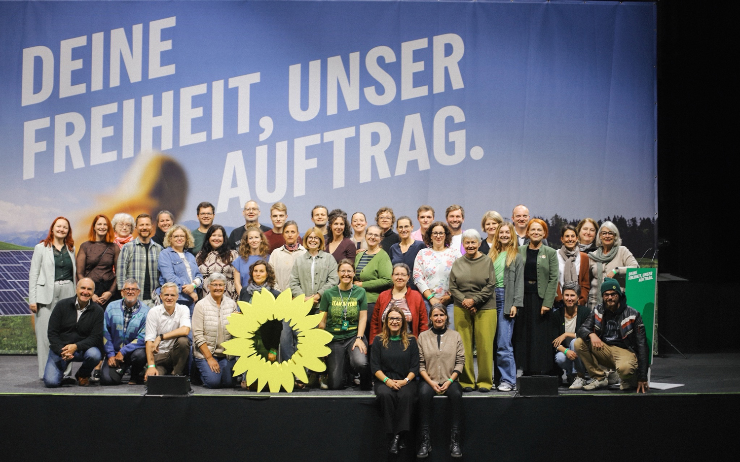 Eine Gruppe von etwa 30 Frauen und Männern posiert gemeinsam auf einer Bühne. Sie stehen und hocken in mehreren Reihen vor einem großen blauen Hintergrundbild, auf dem in weißen Großbuchstaben der Slogan „DEINE FREIHEIT, UNSER AUFTRAG.“ steht. Im unteren Bereich des Bildes, vor der Gruppe, ist eine große, stilisierte gelbe Sonnenblume platziert. Die Personen tragen alltagstaugliche, teils grüne Kleidung und blicken lächelnd in die Kamera.