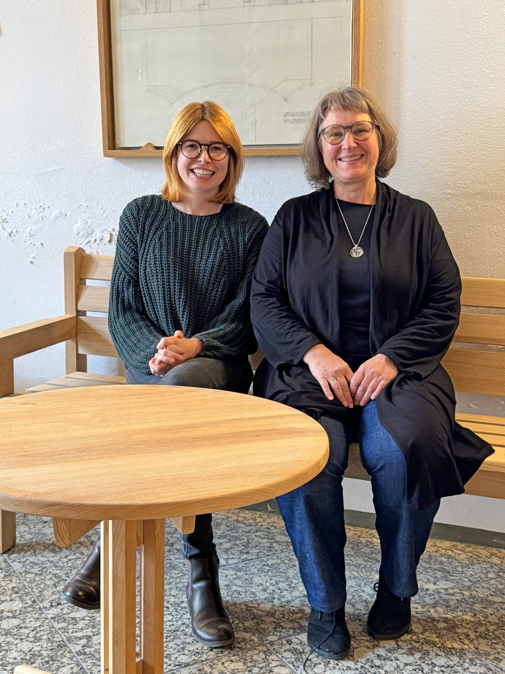 2 Damen sitzen auf einer Holzbank in einem Wirtshaus und lachen in die Kamera.