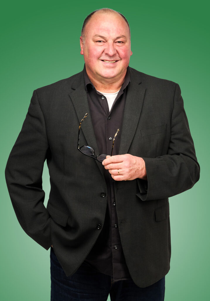 Mann im schwarzem Anzug hat seine Brille in der Hand und lächelt in die Kamera.