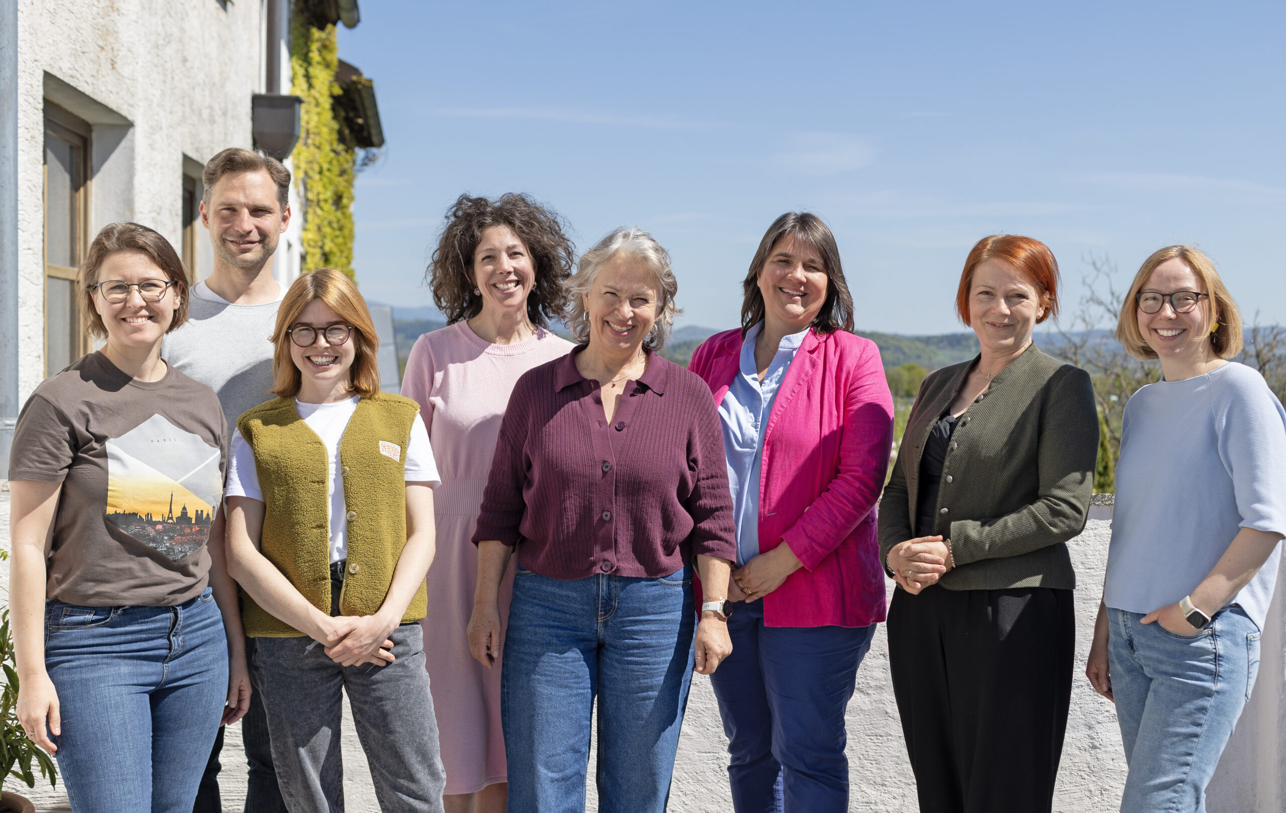 7 Frauen und 1 Mann stehen auf einer Terrasse und blicken lächelnd in die Kamera, Im Hintergrund blauer Himmel und hügelige Landschaft.