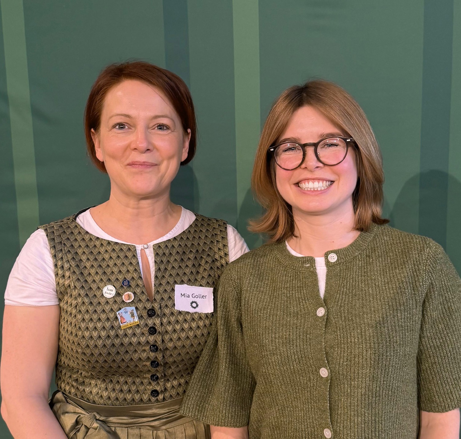 Porträtfoto von zwei lächelnden Frauen vor einem grün gestreiften Hintergrund. Links eine Frau in traditioneller Tracht mit Namensschild „Mia Goller“, rechts eine Frau mit Brille in einem olivgrünen Cardigan.